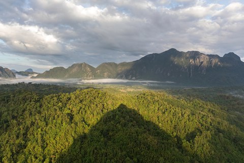 Conical shadow of Mount Nam Xay over green trees at golden hour, South West view from the top, Vang Vieng, Laos